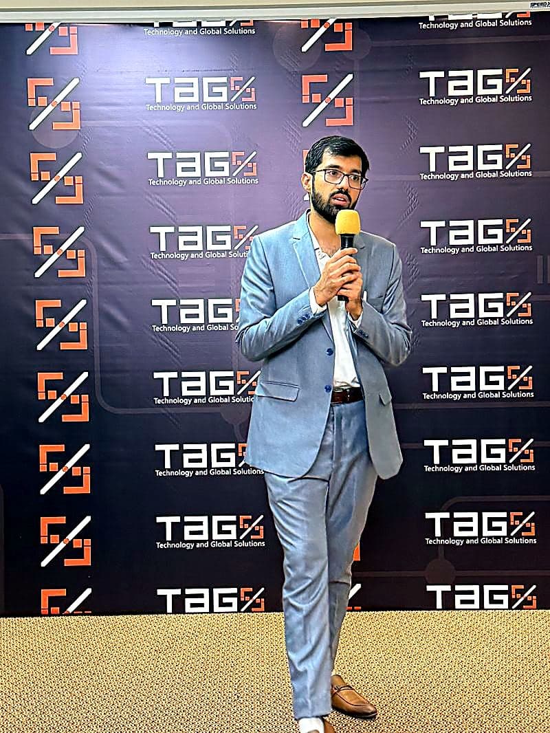 Gohar Naveed speaking with a microphone in front of a TAG Solutions backdrop